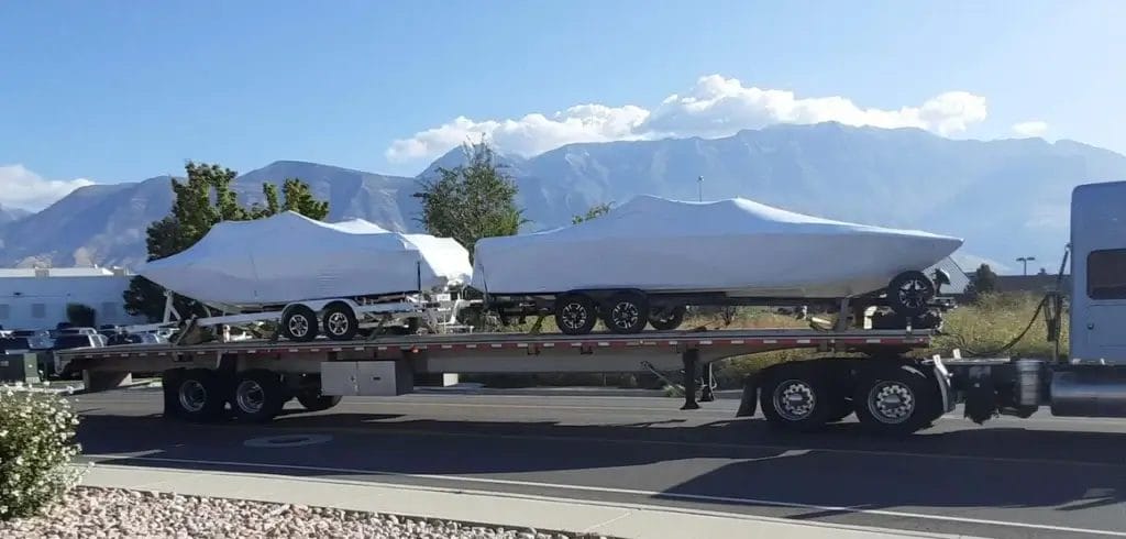Flatbed truck transporting two covered boats on trailers with mountains in the background